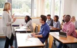 Dozentin spricht vor einer Gruppe Erwachsener in einem modernen Seminarraum. Die Teilnehmerinnen und Teilnehmer, unterschiedlicher Herkunft und Altersgruppen, sitzen an Tischen mit Laptops und Schreibmaterialien und hören aufmerksam zu. Das Klassenzimmer ist hell und freundlich mit großen Fenstern.