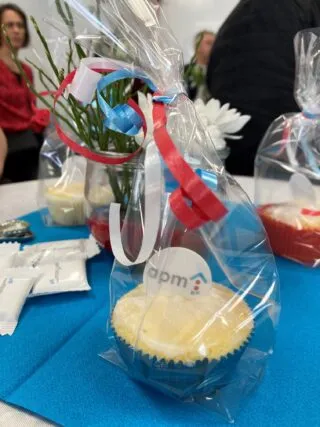 Ein verpackter Muffin mit apm-Logo als Dekoration auf einem gedeckten Tisch, umgeben von weiteren Gastgeschenken, Servietten und kleinen Blumengestecken. Die Schleife in Rot, Weiß und Blau greift die Farben des apm-Logos auf. Im Hintergrund sitzen Menschen an einem Tisch.