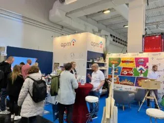 Messestand der apm auf einer Karrieremesse. Besucher:innen informieren sich im Gespräch mit dem apm-Team an einem Stehtisch. Der Stand ist mit dem apm-Logo, Infomaterialien und farbenfrohen Comic-Motiven zum Thema Ausbildung und Karriere gestaltet