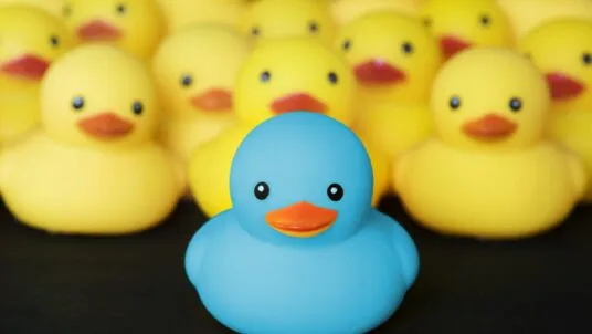 Eine auffällige blaue Quietscheente steht im Vordergrund, umgeben von vielen gelben Quietscheenten im Hintergrund. Symbolbild für Individualität und Anderssein