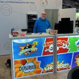 Mann am apm-Messestand mit beleuchtetem Tresen im Comic-Stil. Auf der Theke bunte Begriffe wie ‚Leidenschaft‘, ‚Teamwork‘, ‚Pflege‘ und ‚Karriere!‘. Im Hintergrund das apm-Logo und der Slogan ‚Bildung aus einer Hand – Aus- und Weiterbildung für dein Team