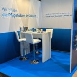 Messestand mit blau-weißer Farbgestaltung und Slogan 'Wir bilden die Pflegehelden der Zukunft' an der Wand. Der Stand verfügt über einen hohen Tisch und zwei weiße Barhocker. Auf dem Tisch liegen Informationsmaterialien, Becher und Schreibutensilien. Die Umgebung ist hell ausgeleuchtet, der Boden ist in Blau gehalten