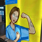 Gelber Hintergrund mit Comic-Illustration einer jungen Frau im blauen Pflegekittel, die selbstbewusst den Arm beugt und eine tätowierte Faust zeigt. Sie trägt eine Brille, rote Lippen und hat Tätowierungen auf der Brust und dem Unterarm. Links oben steht in einem blauen Kasten der Slogan: 'Pflege braucht Mutmacher. Mach deine Ausbildung beim größten privaten Bildungsanbieter für Pflege!' Daneben das Logo der apm mit stilisierten Punkten. Die Szene vermittelt Stärke, Selbstbewusstsein und Individualität von Pflegekräften.