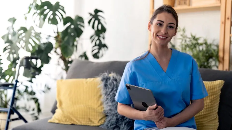 Pflegekraft in blauer Dienstkleidung sitzt mit einem Tablet auf dem Schoß auf einem Sofa. Sie lächelt freundlich in die Kamera. Im Hintergrund stehen grüne Pflanzen, gelbe Sofakissen und ein Rollator – die Szene wirkt wohnlich und modern.